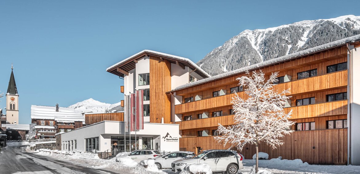 Das Sporthotel Silvretta Montafon von vorne mit Schnee bedeckt und der Kirche und dem Dorf im Hintergrund mit Berge | © Silvretta Montafon - blateral