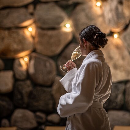 Gold Spa im Sporthotel Silvretta Montafon | © Silvretta Montafon - Mara Baues Eine Frau in einem Bademantel trinkt einen Prossecco im Tepidarium mit goldenem Licht in den Steinwänden | © Silvretta Montafon - Mara Baues