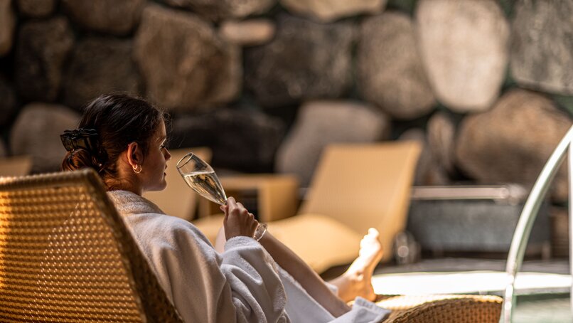 Gold Spa im Sporthotel Silvretta Montafon | © Silvretta Montafon - Mara Baues Eine Frau in einem Bademantel trinkt einen Prossecco im Tepidarium auf einer Liege | © Silvretta Montafon - Mara Baues